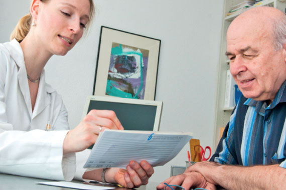 Eine Ärztin in einem weißen Kittel erklärt einemälteren Patienten seine Werte auf einem Blatt Papier.